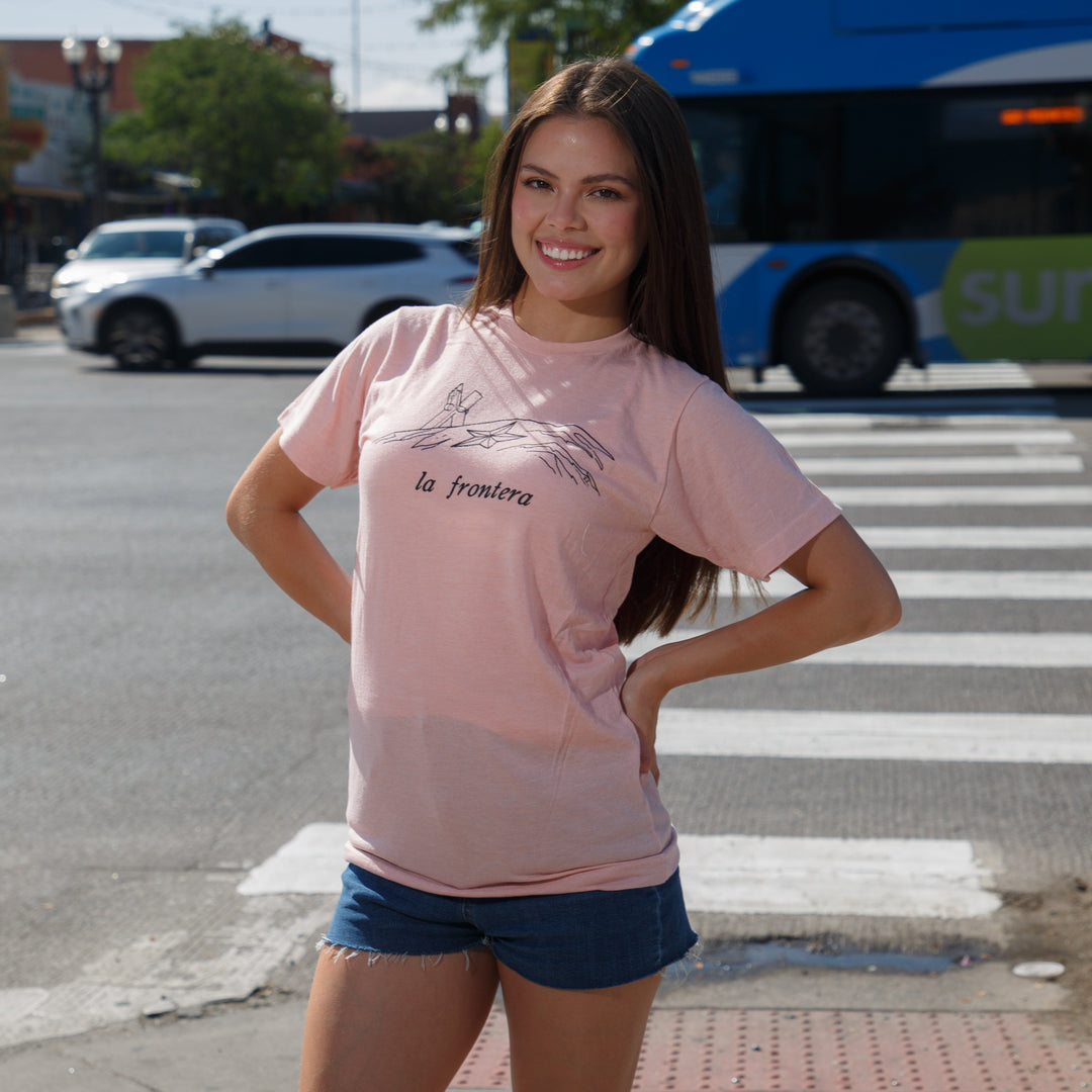 Woman in a pink shirt and blue shorts standing on a city street corner with traffic lights and a bus in the background.
